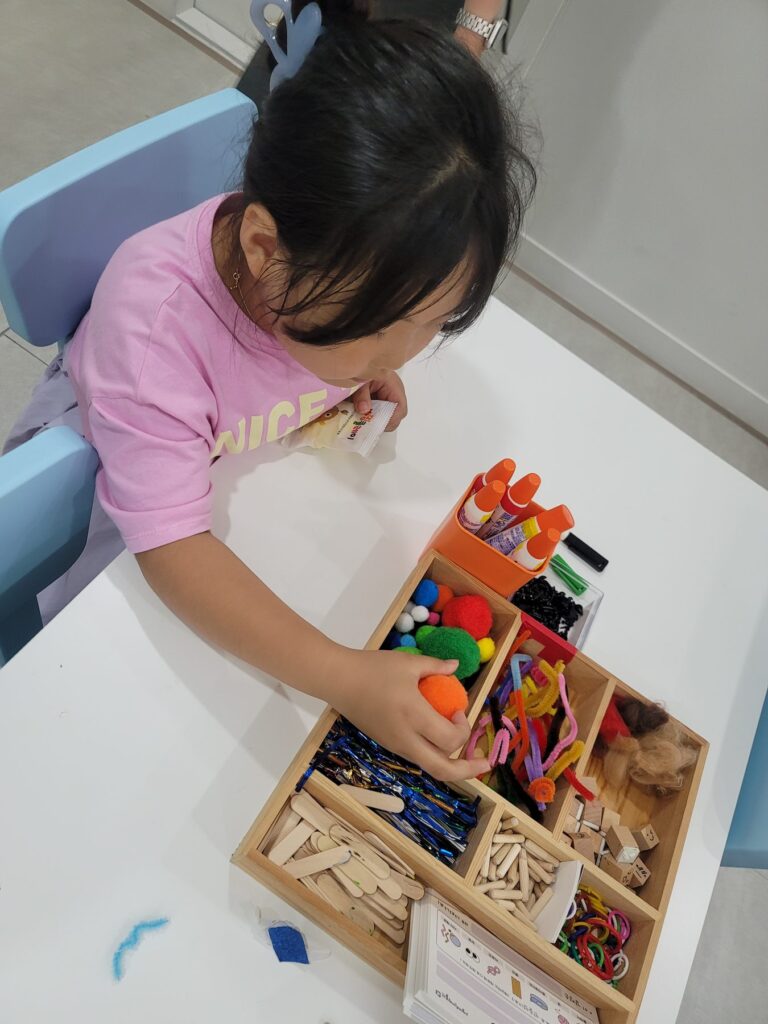Child choosing pom-poms and pipe cleaners from a wooden craft tray