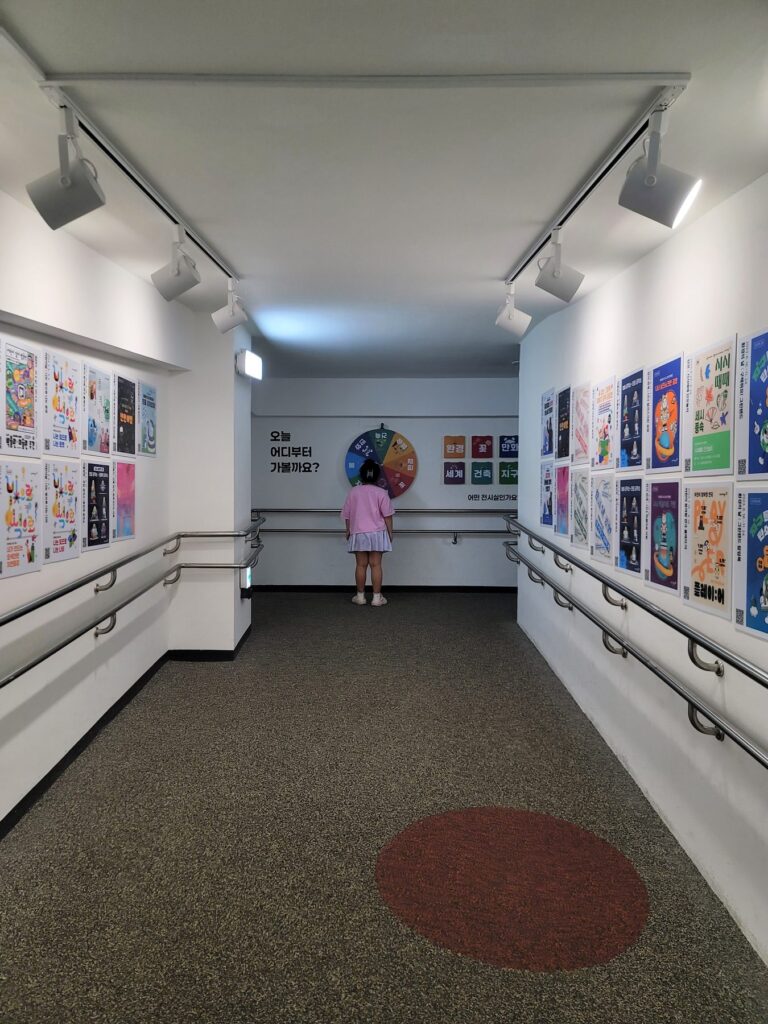 Poster-lined corridor at Goyang Children's Museum with a spinner asking which exhibition hall to visit first